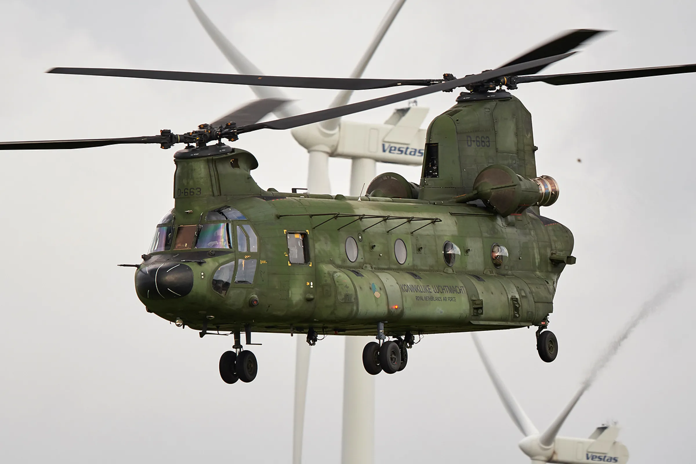 Een Chinook helikopter van Defensie vliegt voor twee windturbines langs.